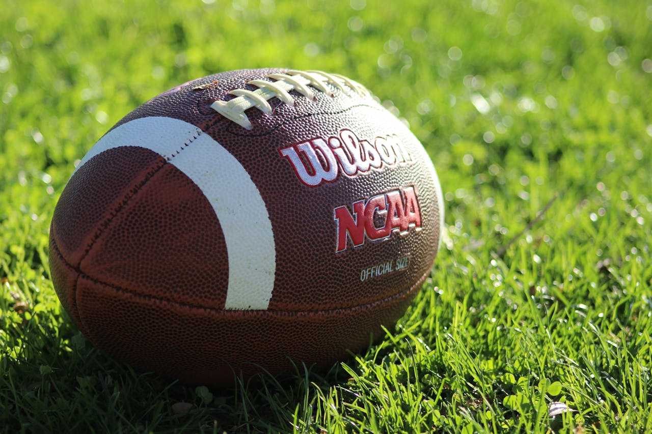 Detailed image of a Wilson NCAA football resting on vibrant green grass in daylight.